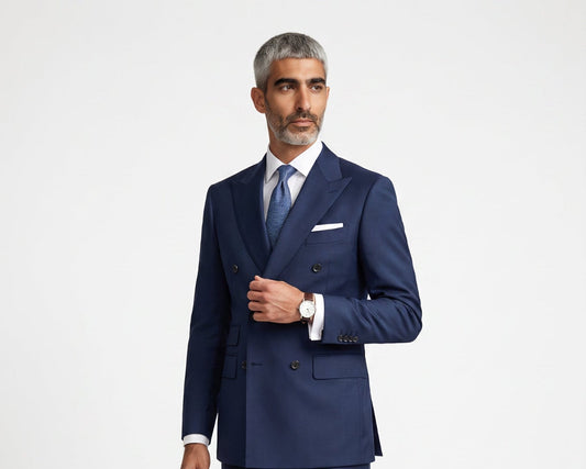 A man modeling a navy blue double-breasted suit with a white shirt, blue tie, and white pocket square, standing against a light gray studio background.