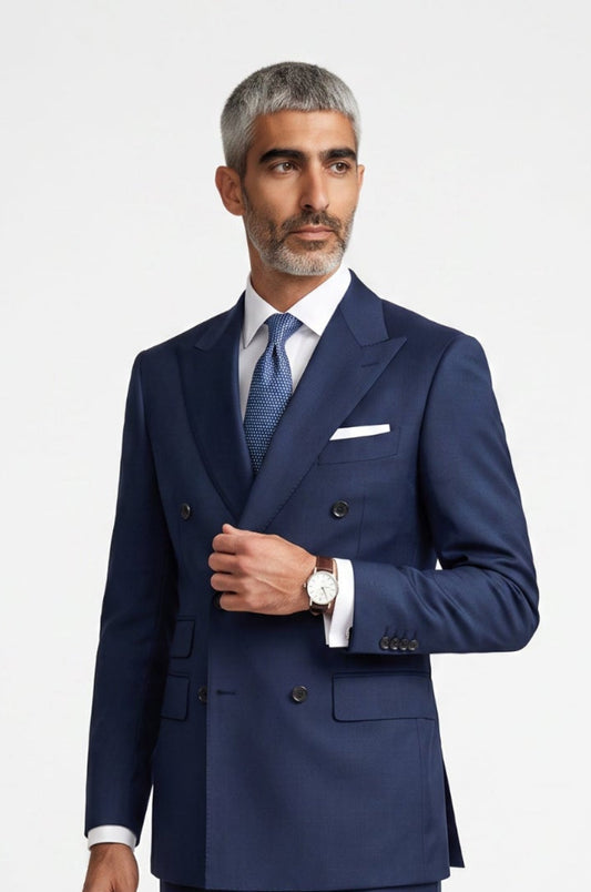 A man modeling a navy blue double-breasted suit with a white shirt, blue tie, and white pocket square, standing against a light gray studio background.