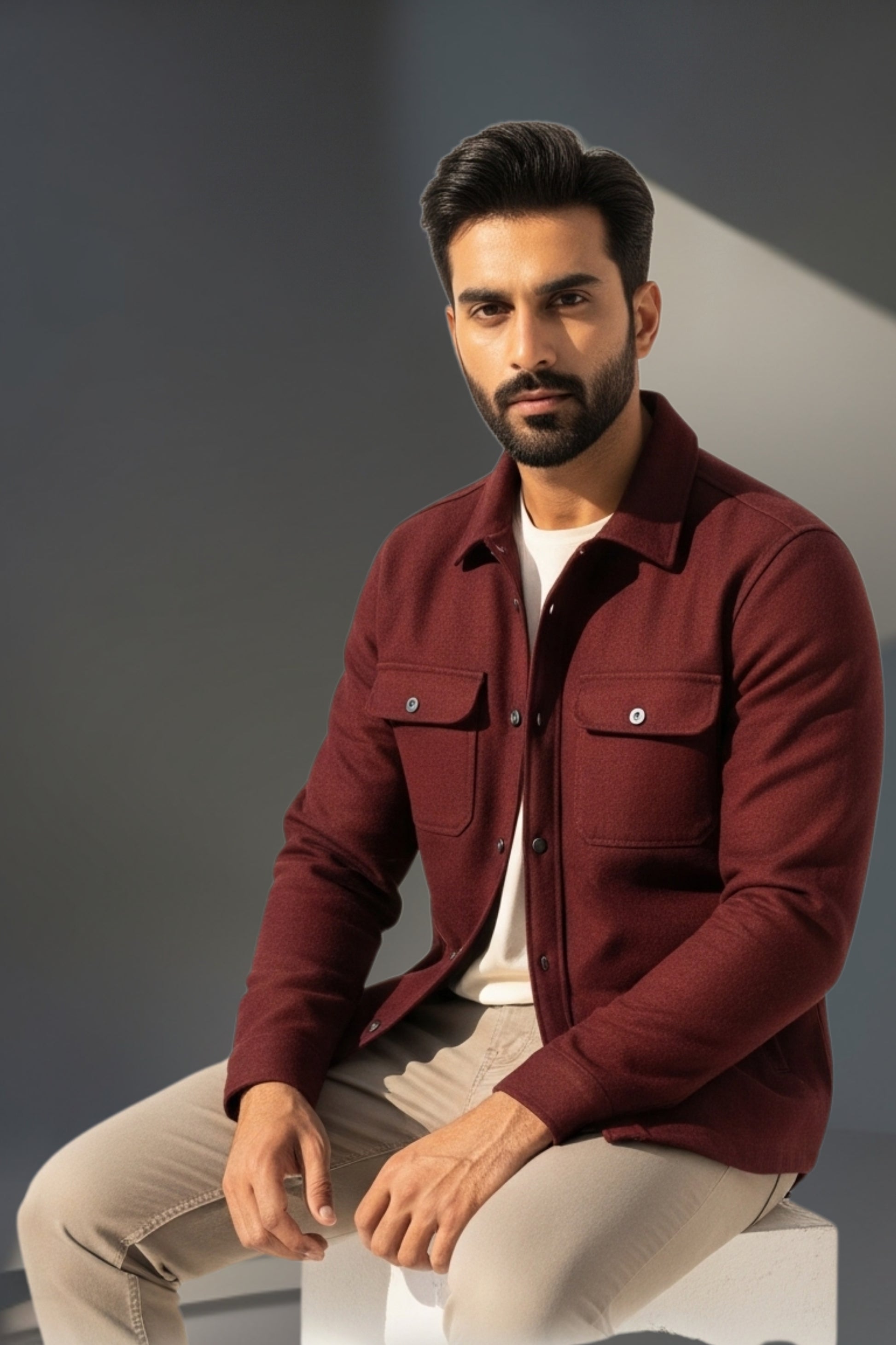 Man wearing a maroon jacket sitting on a white stool against a gray background