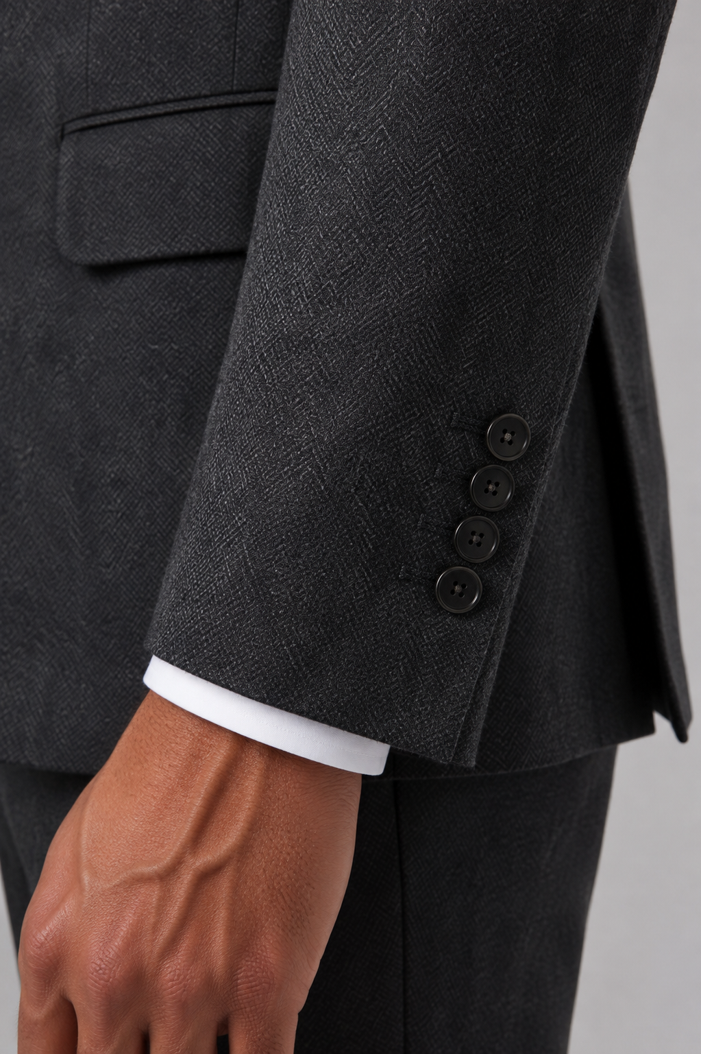 Close-up of a dark charcoal gray herringbone suit jacket sleeve with four buttons, worn over a white dress shirt cuff, on a mannequin or model.