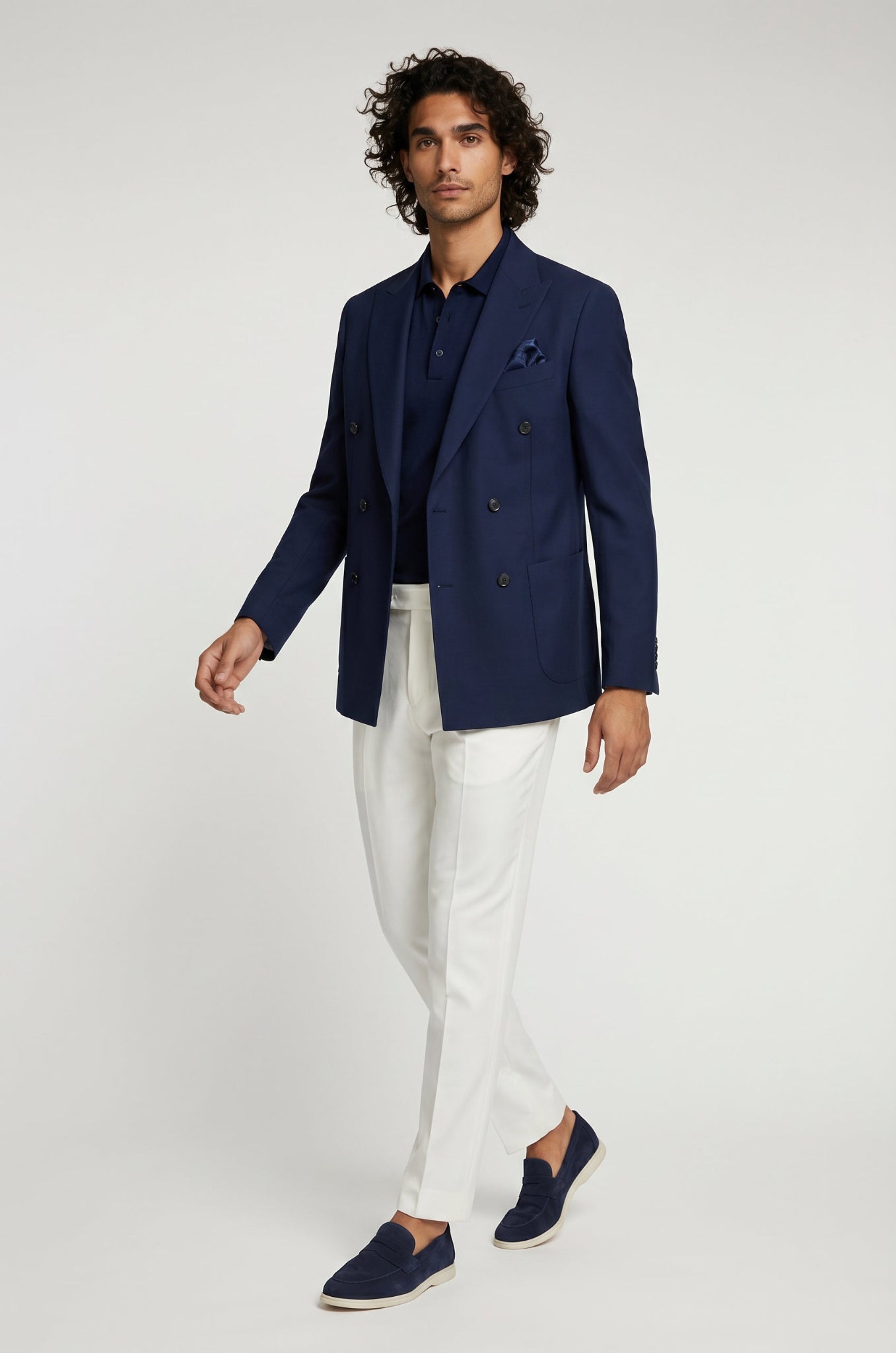 Man wearing a navy blazer, white pants, and navy shoes on a light gray background