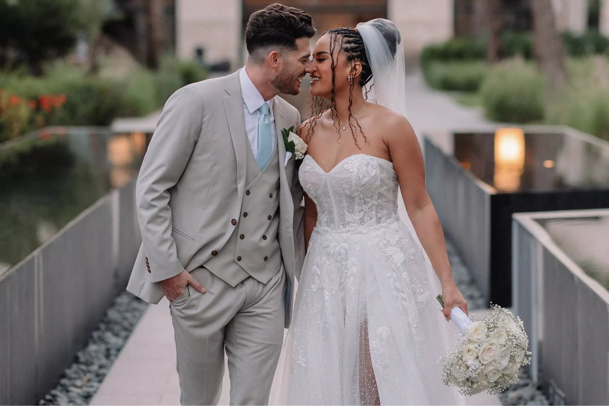 A bride in a white lace wedding gown and veil walking beside a groom in a light gray double-breasted suit with a light blue tie, holding a bouquet in an outdoor garden setting with palm trees and a modern walkway.