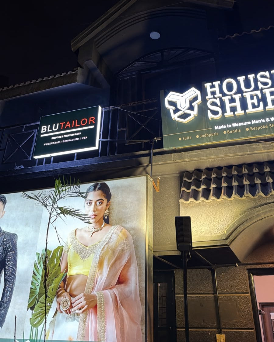 Nighttime exterior of a building with illuminated signs reading HOUSE SHEF and BLUTAILOR, plus a large fashion poster showing a woman in a light pink outfit.