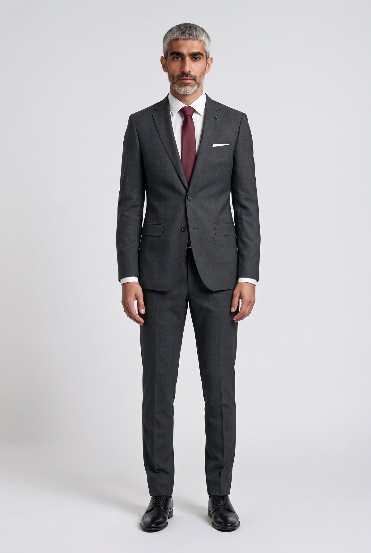 A man wearing a dark gray two-piece suit with a white dress shirt, a burgundy tie, and black dress shoes, standing against a light gray studio background.