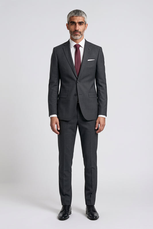 A man wearing a dark gray two-piece suit with a white dress shirt, a burgundy tie, and black dress shoes, standing against a light gray studio background.